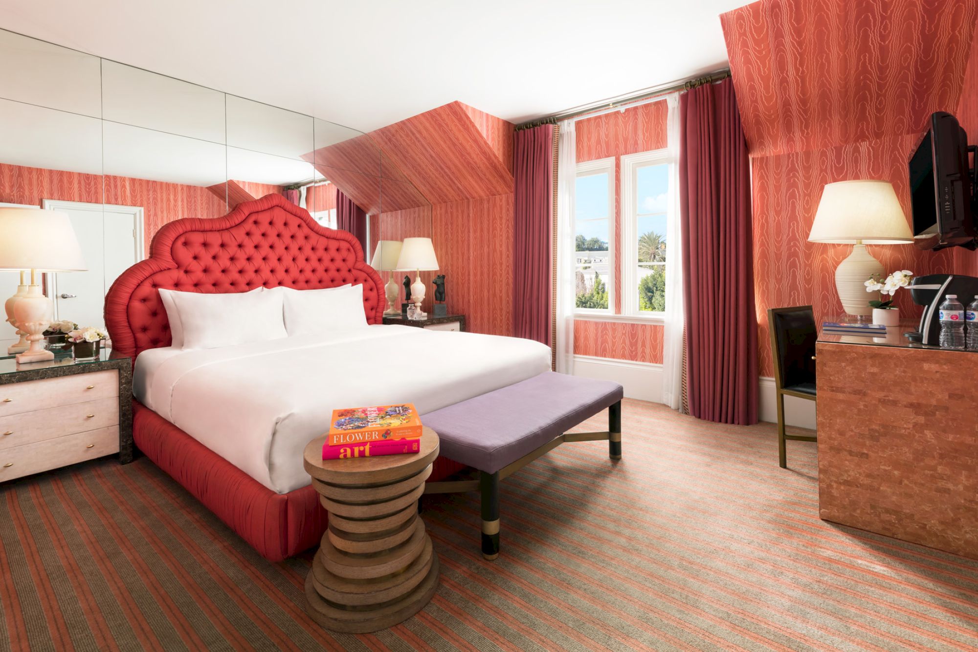 A stylish hotel room with a red tufted headboard, lamps, mirrored wall, desk, and decorative accents.