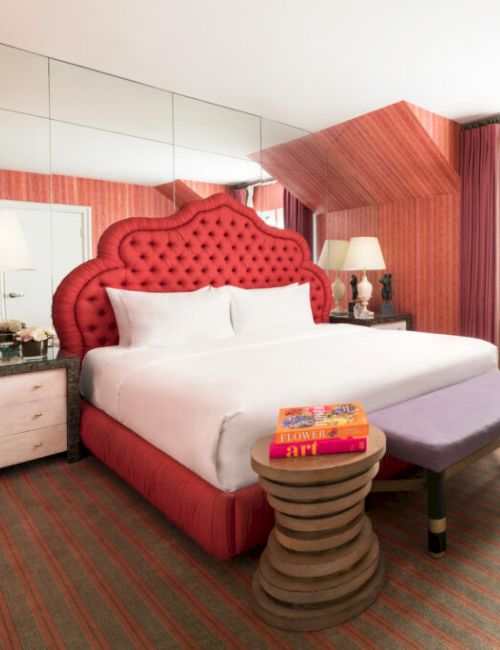 A stylish hotel room features a bold red bed, mirrored wall, lamps, books, chair, and a view through large windows with red curtains.