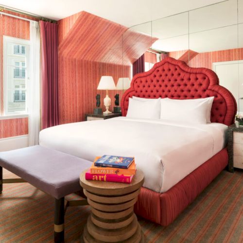 Elegantly decorated hotel room with a large bed, vibrant headboard, bedside lamps, and a stack of books on a table, exuding cozy comfort.