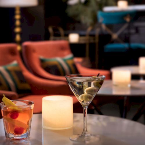 A cozy lounge setting with two cocktails on a table, one in a rocks glass with garnish, and another in a martini glass with olives.