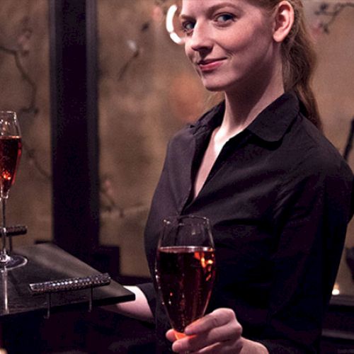 A person in a black outfit holds a tray with two glasses of red liquid in an elegant room with decorative wall art behind them.