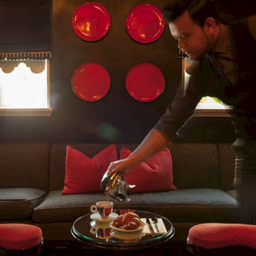 A person is serving coffee and croissants on a table in a cozy room with red decor accents and cushioned seating.