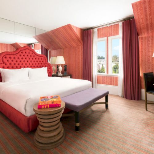 A stylish hotel room with a red tufted bed, matching curtains, a mirrored wall, bedside tables, and a desk with a lamp and chair.