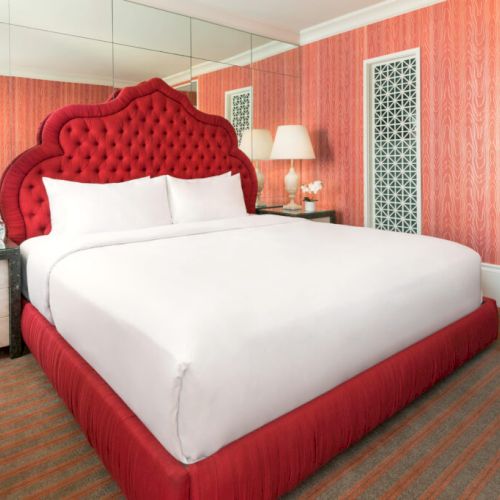 A hotel room with a red tufted headboard, white bedding, mirrored wall, lamps, and patterned wallpaper.