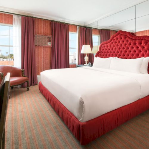 A hotel room with a large red bed, desk, chair, lamps, and art. Curtains cover windows on one side, adding a luxurious touch.