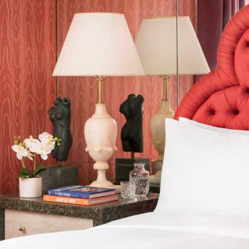 The image shows a bedroom with a red tufted headboard, a lamp, books, and a small statue on a bedside table, with a pink textured wall.