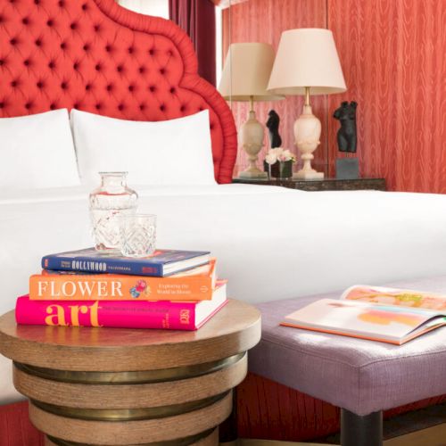 A cozy bedroom scene with a red tufted headboard, books, a bedside lamp, and a round wooden table.