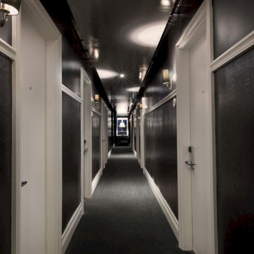 A dimly lit hotel corridor with black walls, white doors, room numbers visible, and a framed picture at the hall's end.