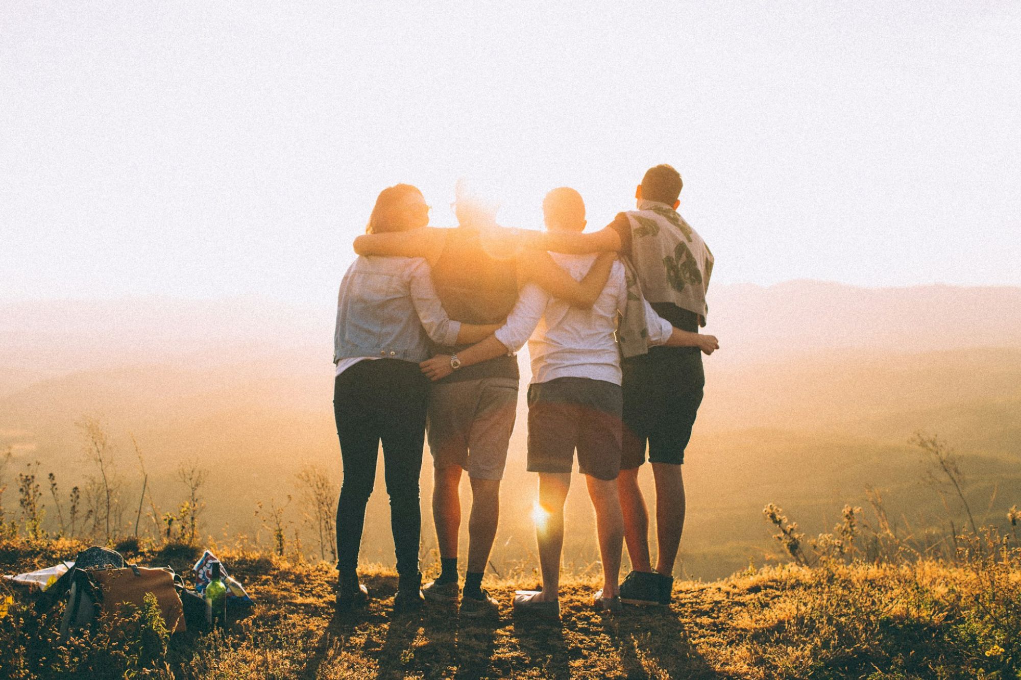 Four people stand together, embracing while watching a sunset over a scenic landscape, with a bag and bottle nearby.