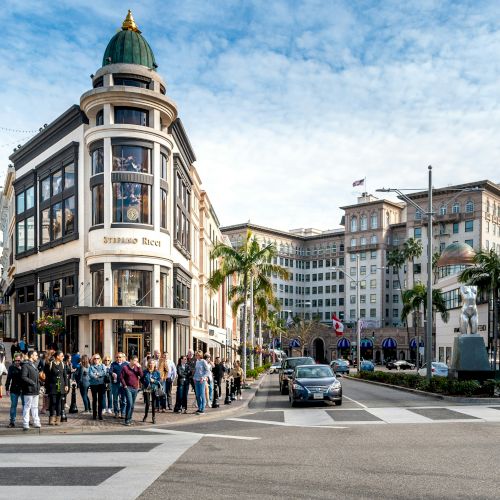 A bustling city street corner with a domed building, palm trees, cars, and pedestrians crossing a road under a clear sky.