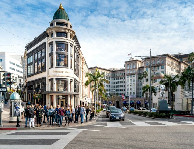 A bustling city street corner with a domed building, palm trees, cars, and pedestrians crossing a road under a clear sky.